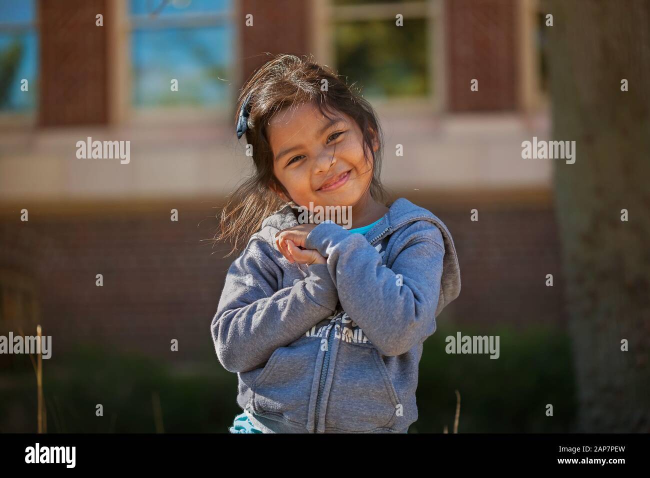 Süßes kleines Mädchen, das ihre Hände zusammenhält, die Liebe, Glück oder Gratis-Begabung zum Ausdruck bringt, mit einem Schulgebäude aus Backsteinen im Hintergrund. Stockfoto