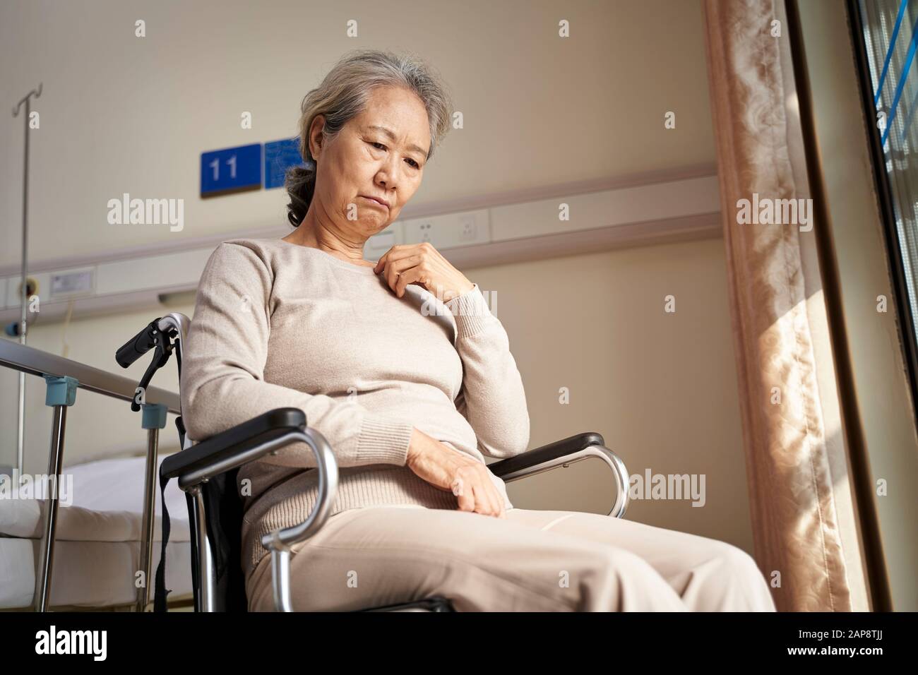 Traurige und depressive asiatische ältere Frau, die allein im Rollstuhl sitzt und im Pflegeheim mit dem Kopf nach unten steht Stockfoto