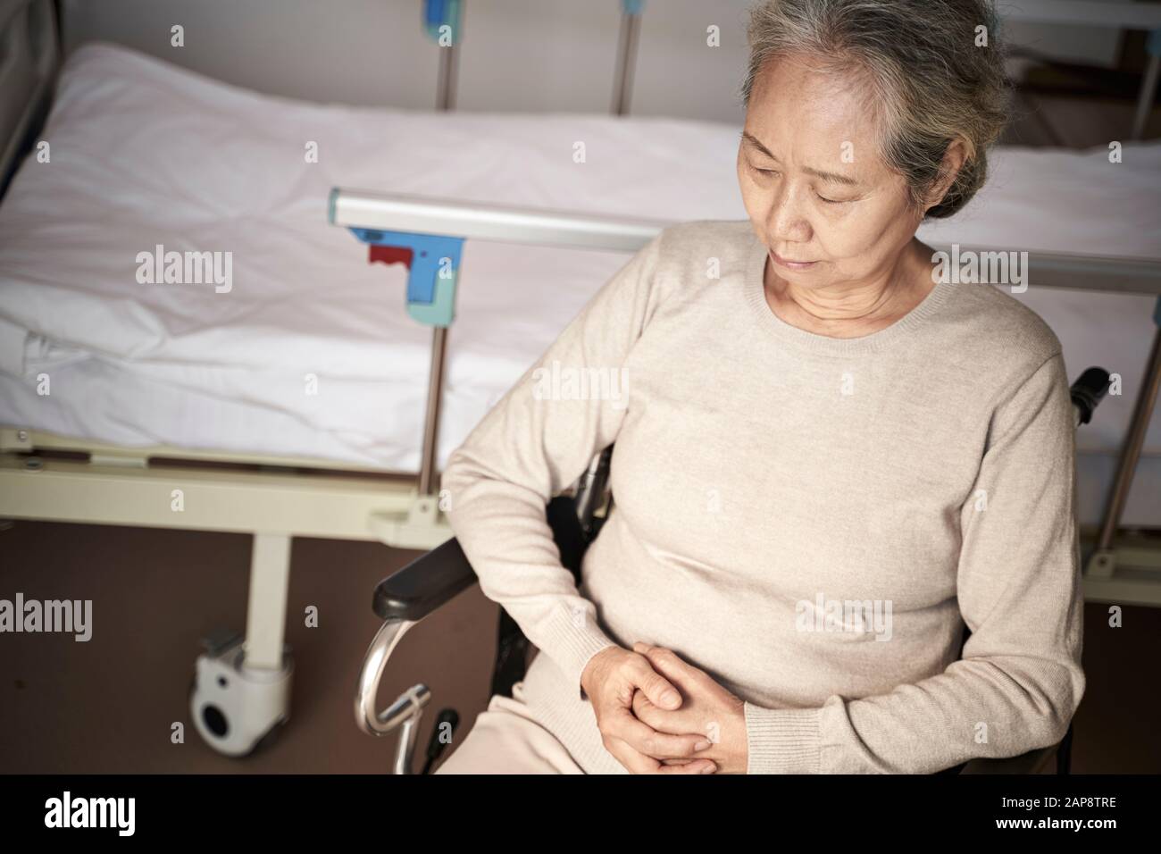 Hochwinkeliger Blick auf die traurige asiatische Frau, die im Rollstuhl neben dem Krankenhausbett sitzt Stockfoto