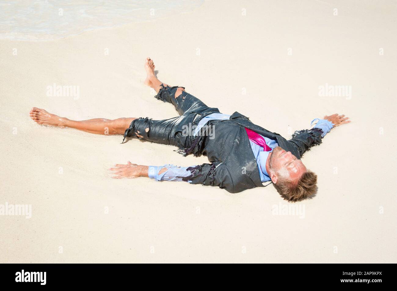 Castaway Survivor Geschäftsmann, der am Ufer eines tropischen Strandes in einem zerrissenen Anzug liegt Stockfoto