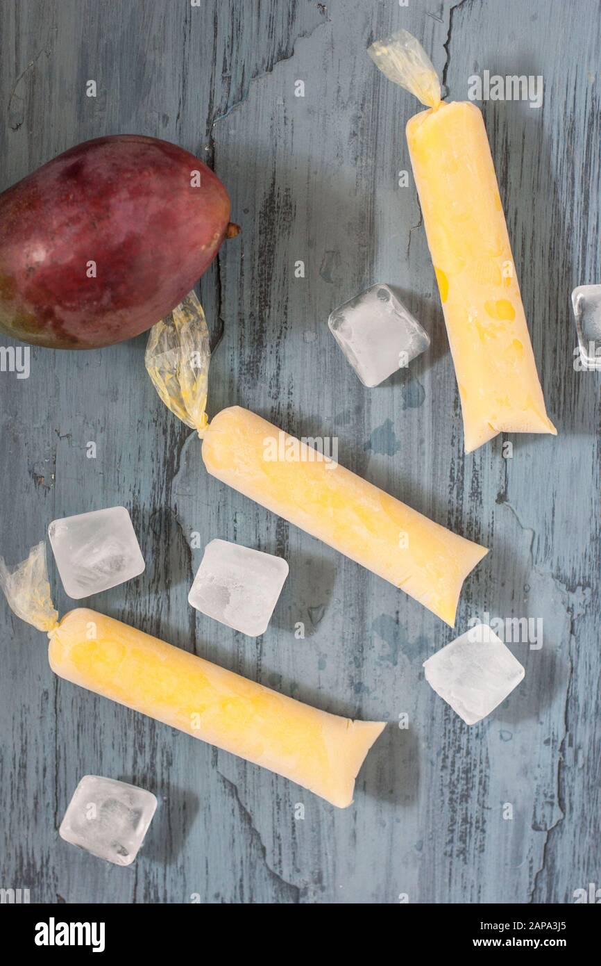 Drei cremige Mango-Lollies auf blauem Grund mit ganzen Mango-Früchten und Eiswürfeln Stockfoto