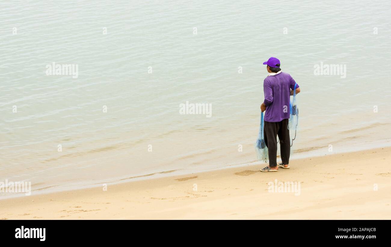 Thailändischer Fischer am Wasserrand mit blauem Netz bei Ebbe mit lilafarbenem Hut und Hemd mit blauem Netz Stockfoto