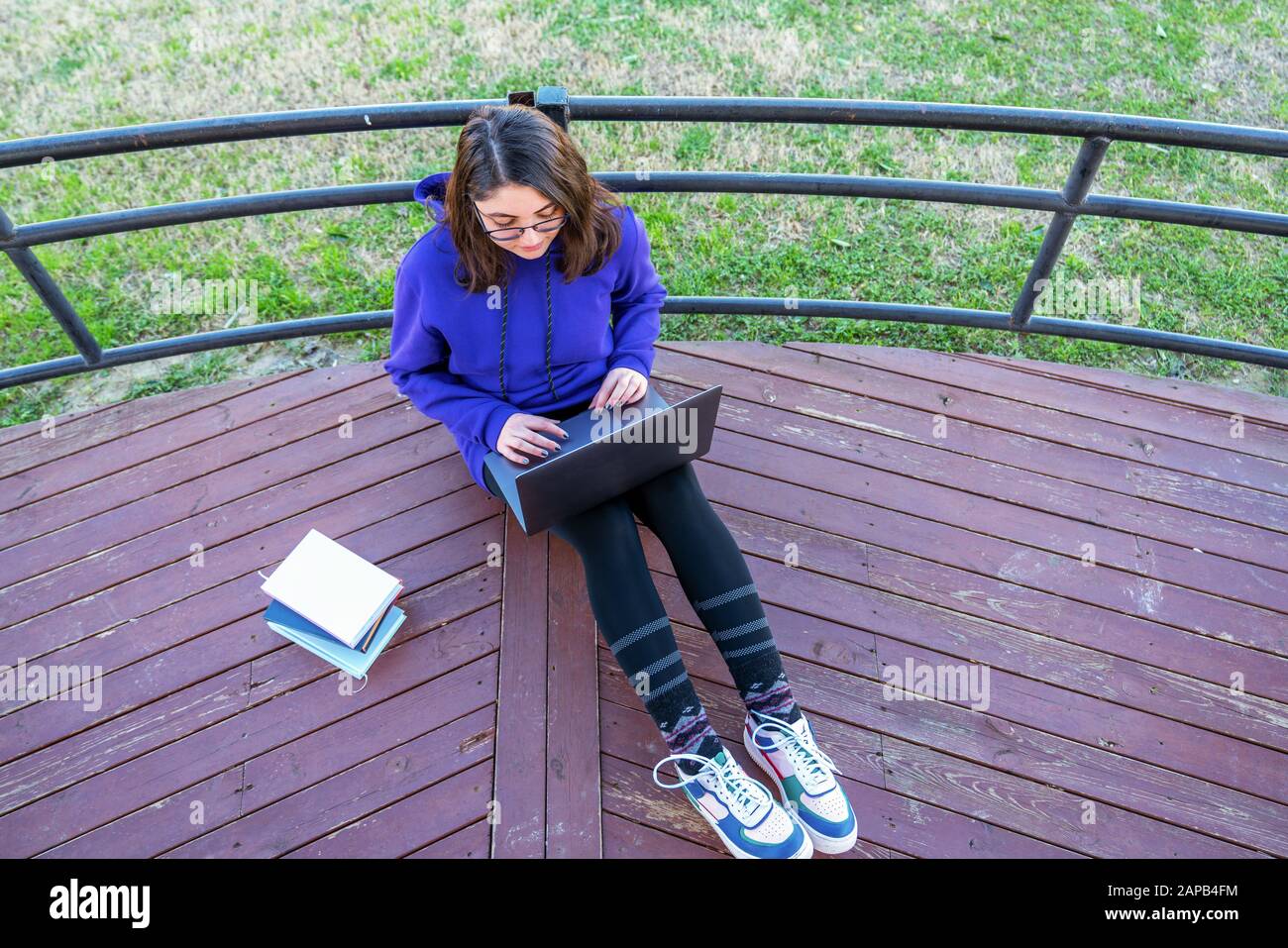Intellektuelles junges Mädchen sitzt auf der Terrasse und benutzt einen Laptop mit Büchern neben ihr in einem Park Stockfoto