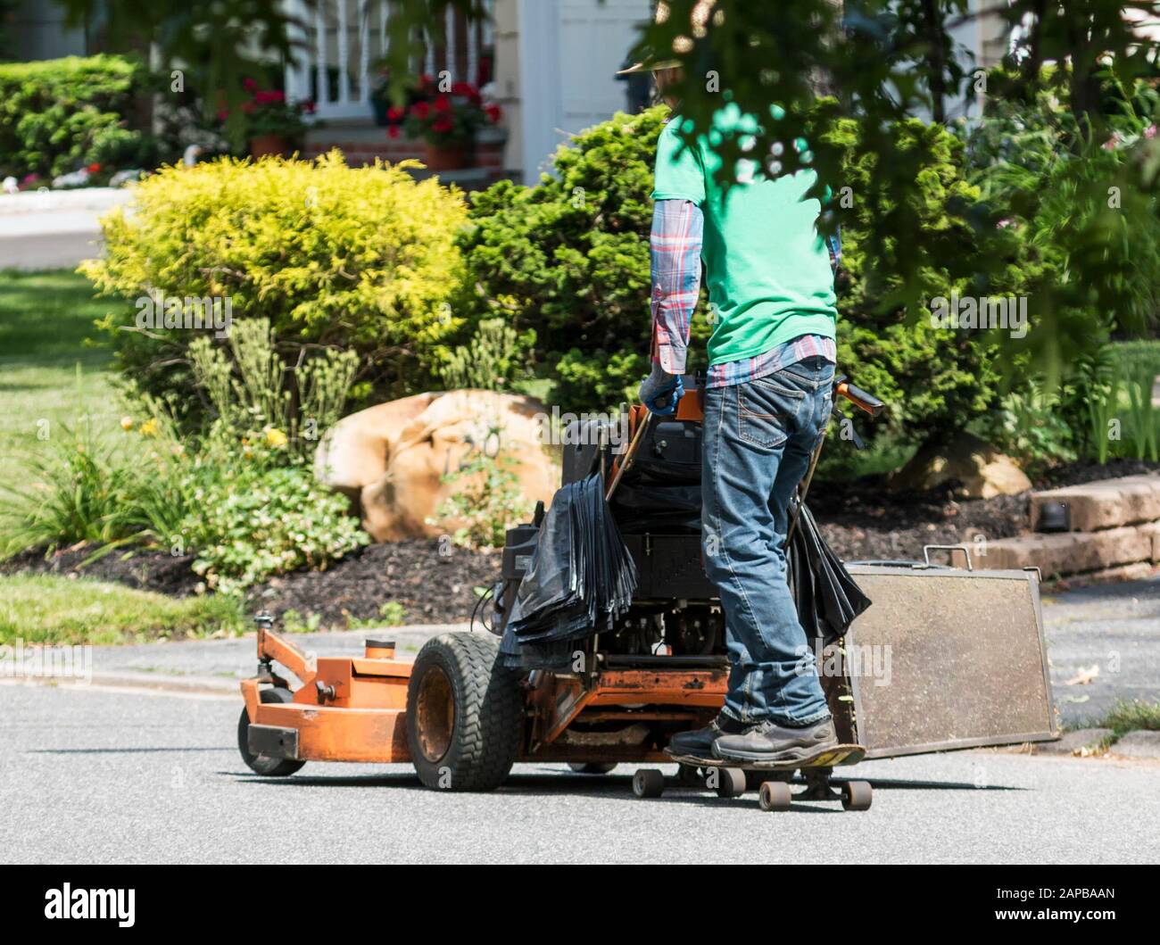 Ein Landschaftsgestalter kommt zu einem anderen Job, indem er hinter seinem Rasenmäher mit einem Skateboard auf der Straße fährt. Stockfoto