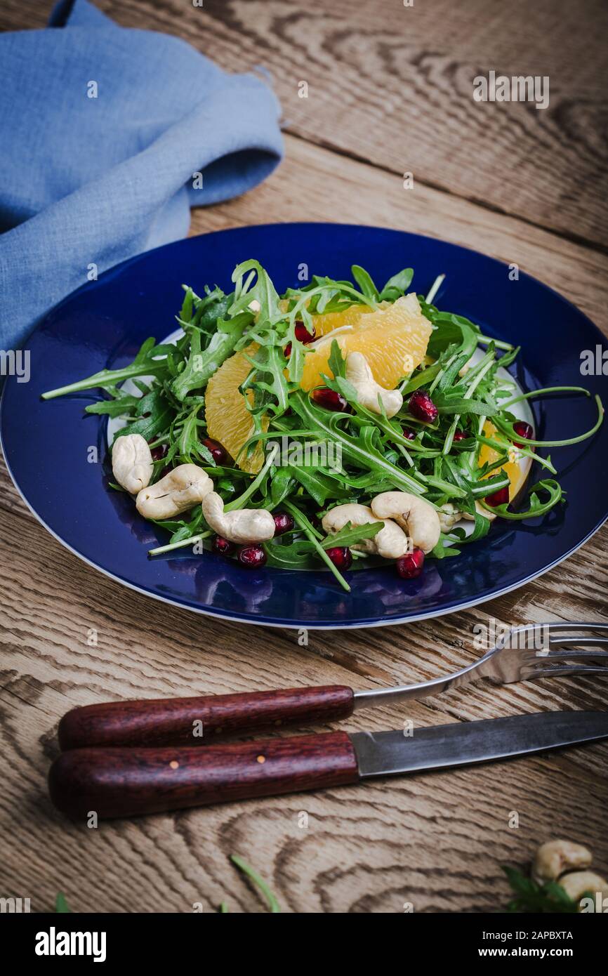 Veganer Salat auf Pflanzenbasis mit frischem Blattgemüse, Arugula, Orangenscheiben, Granatapfelsamen, Cashews in blauer Schüssel auf rustikalem Holztisch, gesundes li Stockfoto