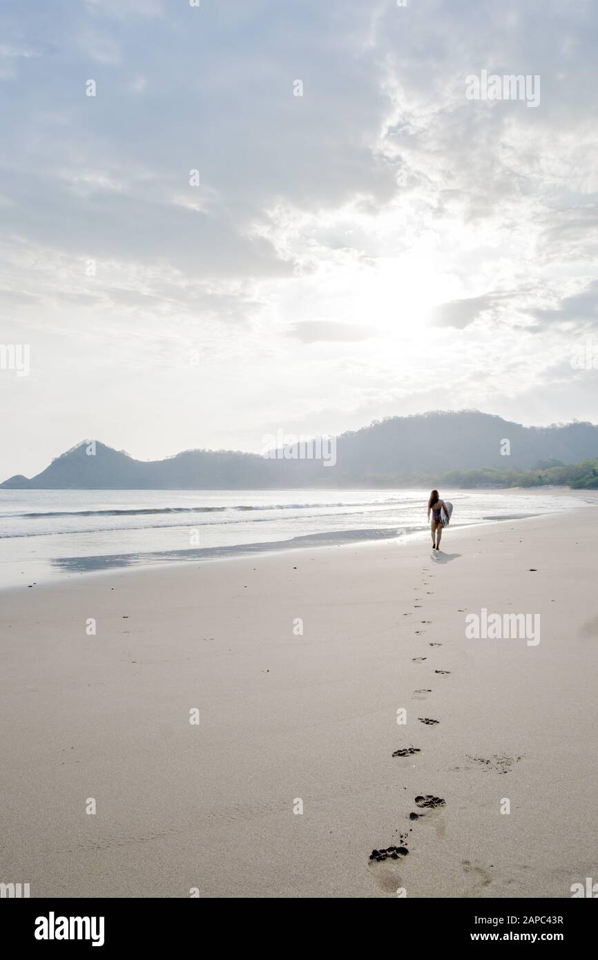 Mittelamerika, Nicaragua, San Juan del Sur. Eine Frau surfer, die Fuß verlässt, fällt bei Sonnenuntergang auf einen leeren weißen Sandstrand Stockfoto