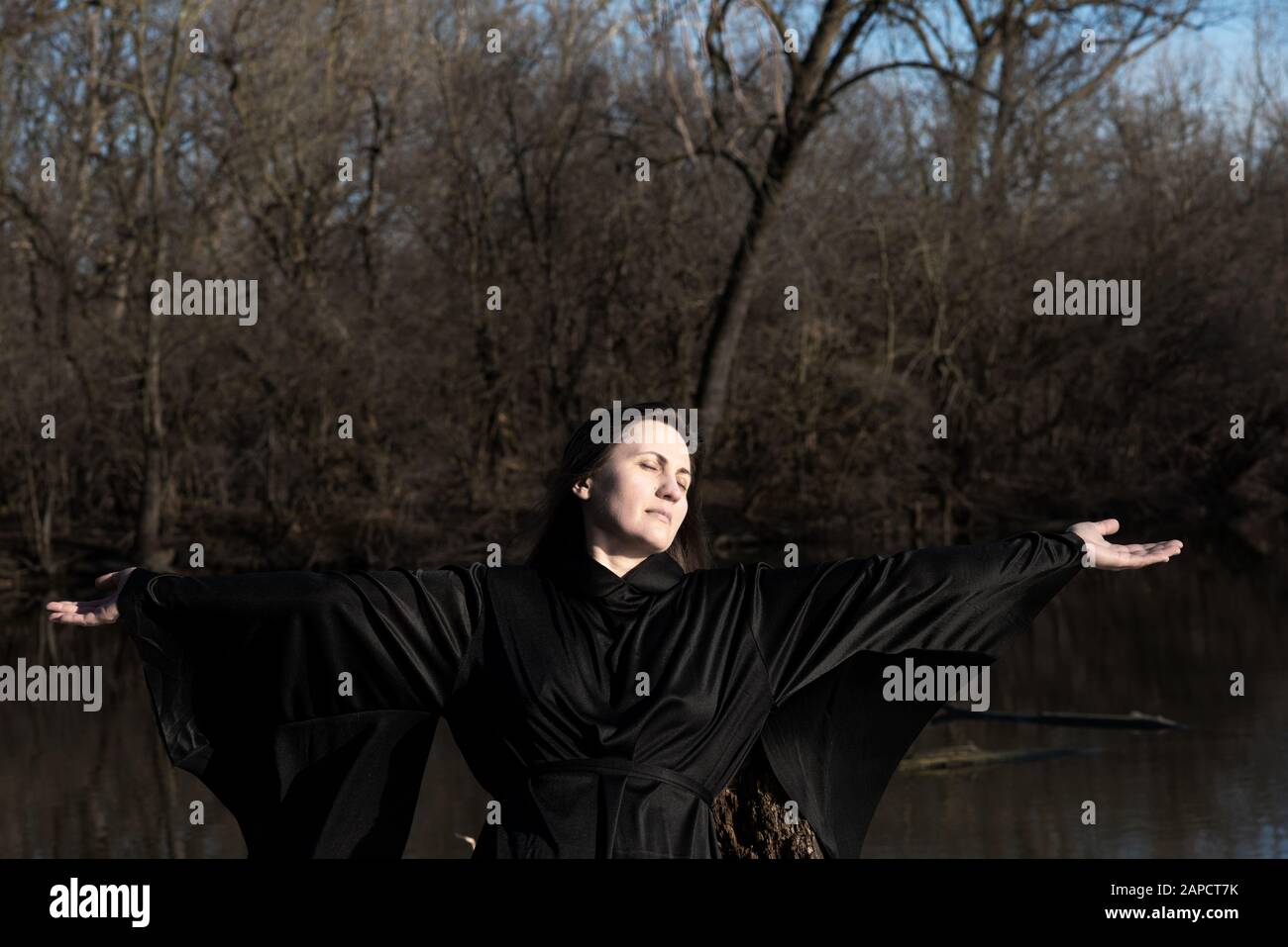 Frau mit dunkellangem Haar in schwarzen Gewändern vor dem See. Magie und Hexerei. Zurück zum Naturkonzept. Stockfoto