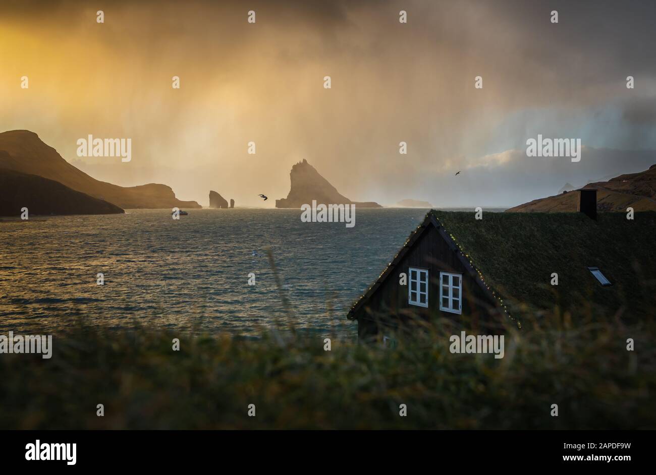 In Richtung der Rasenhäuser im Dorf Bøur zieht eine Schneedusche. Im Hintergrund werden Drangarnir und Tindhólmur von der Sonne beleuchtet. Faröer Stockfoto