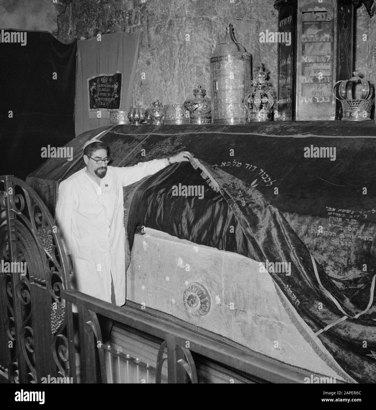 Israel 1964-1965: Jerusalem (Jerusalem), Kirchen und Klöster Beschreibung: Am Grabstein hebt der mutmaßliche Vormund/Gastgeber die Samtoberfläche, um eine ornamentierte Anmerkung zu zeigen: Das Grab befindet sich im Erdgeschoss und dient heute als Synagoge und Betraum. Der Grabstein ist mit einem Samttuch bedeckt und darauf Tora-Kronen aus Synagogenkreisen jüdischer Gemeinden, der Grabstein selbst hat Aufschriften und Verzierungen aus der Kreuzfahrerzeit. Die Letzte Abendmahlshalle und das Grab Davids befinden sich im selben Gebäude Datum: 1964 Ort: Israel, Jerusalem, Berg Zion Schlüsselwörter: Gräber, Stockfoto