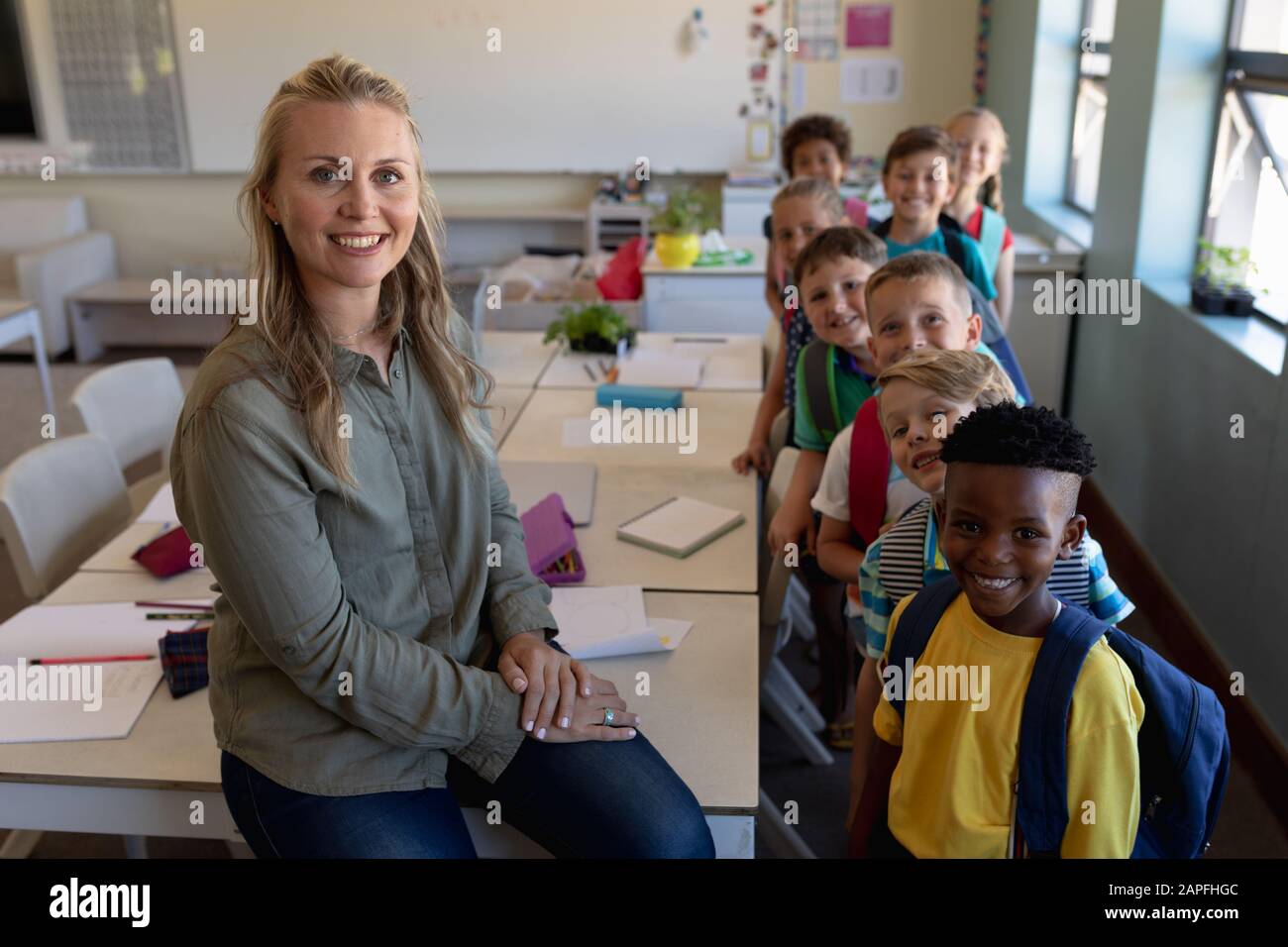 Schullehrerin, die in einem Klassenzimmer mit einer Gruppe von Schulkindern auf einem Schreibtisch sitzt Stockfoto