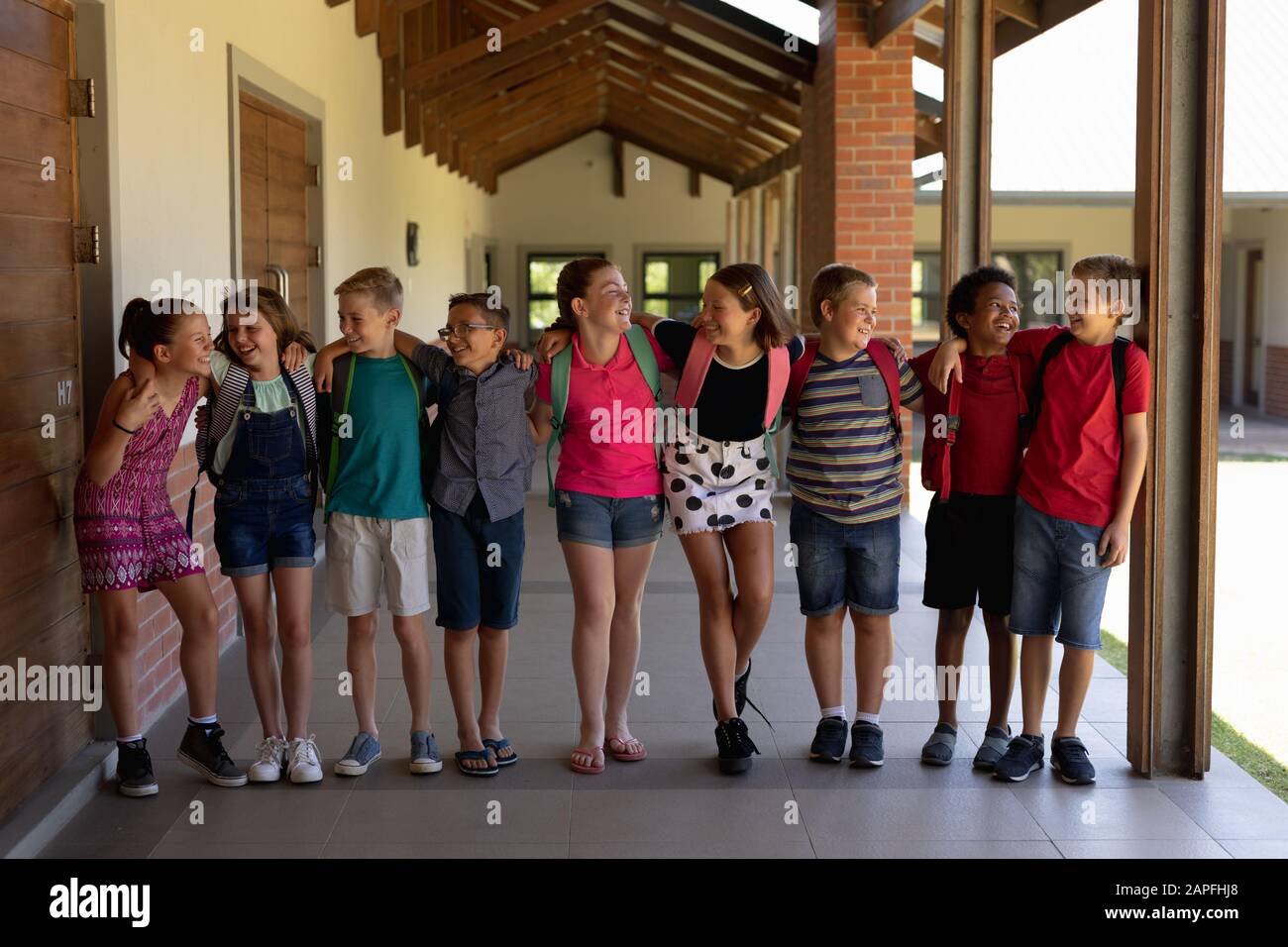 Gruppe von Schulkindern, die in einer Reihe von Armen um sich herum in einem Außenkorridor spazieren Stockfoto