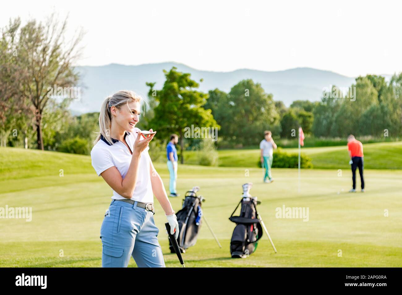 Frau spricht auf dem Smartphone auf dem Golfplatz, Freunde spielen im Hintergrund Golf Stockfoto
