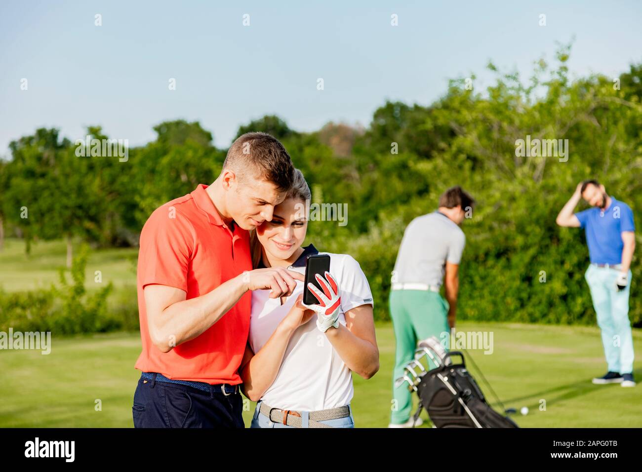 Paare, die Smartphone auf dem Golfplatz nutzen, Freunde, die im Hintergrund Golf spielen Stockfoto
