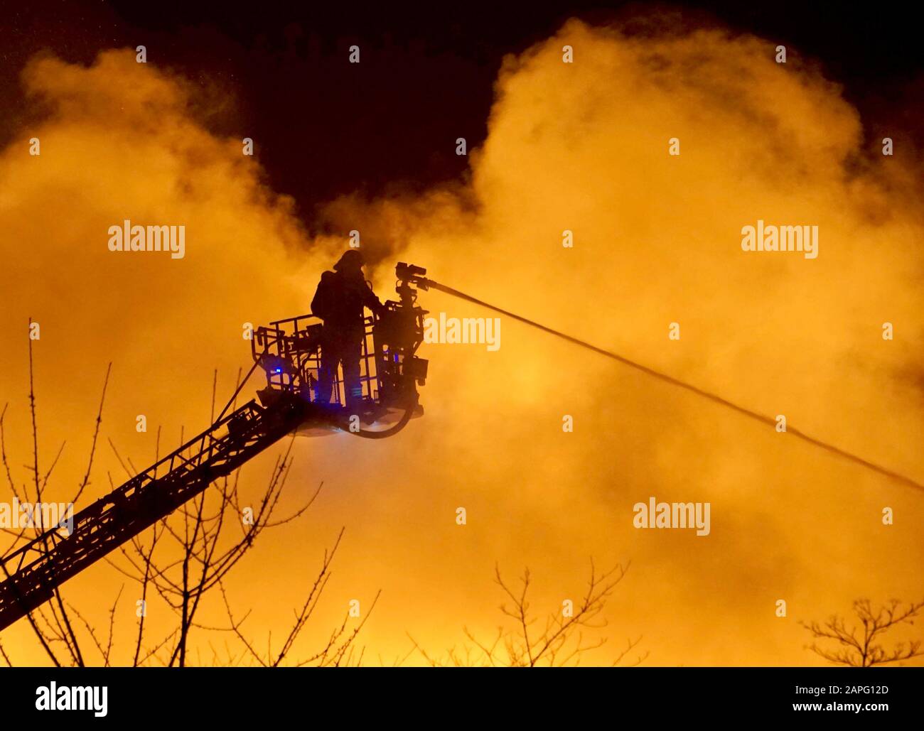 23. Januar 2020, Baden-Württemberg, Ilsfeld: Ein Feuerwehrmann steht auf  einer Drehleiter vor einer Rauchwolke, die während der Löscharbeiten von  Flammen erhellt wird. Das Feuer zerstörte ein Gebäude einer Palettenfirma  im Landkreis Heilbronner