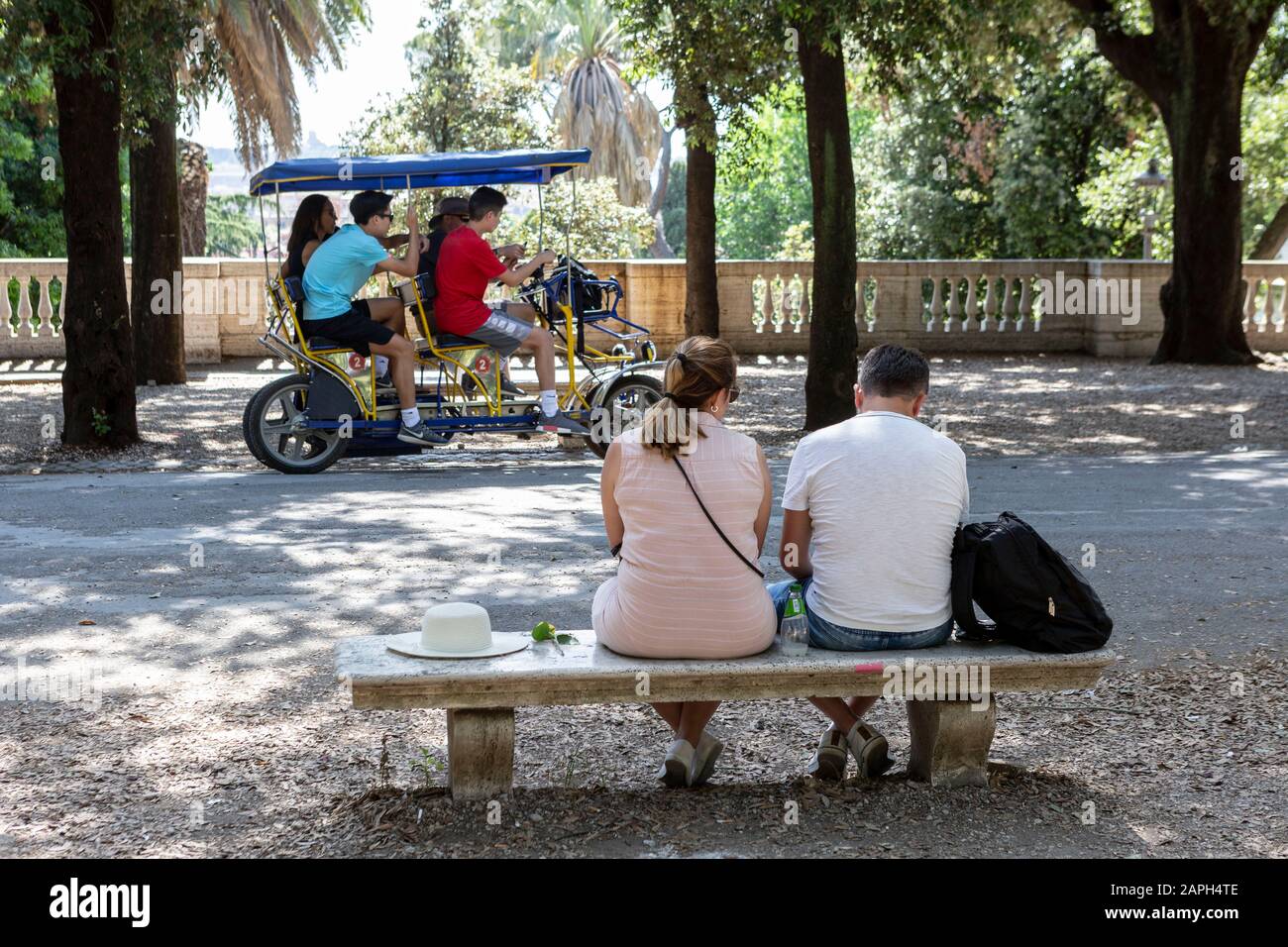 Ein Paar sitzt auf einer Parkbank in der Villa Borghese in Rom, während vier Personen im Hintergrund mit einem Surrey Fahrrad fahren. Stockfoto