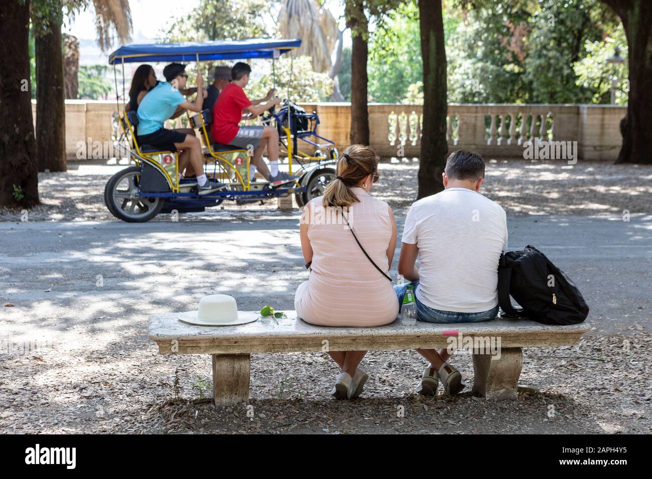 Ein Paar sitzt auf einer Parkbank in der Villa Borghese in Rom, während vier Personen im Hintergrund mit einem Surrey Fahrrad fahren. Stockfoto