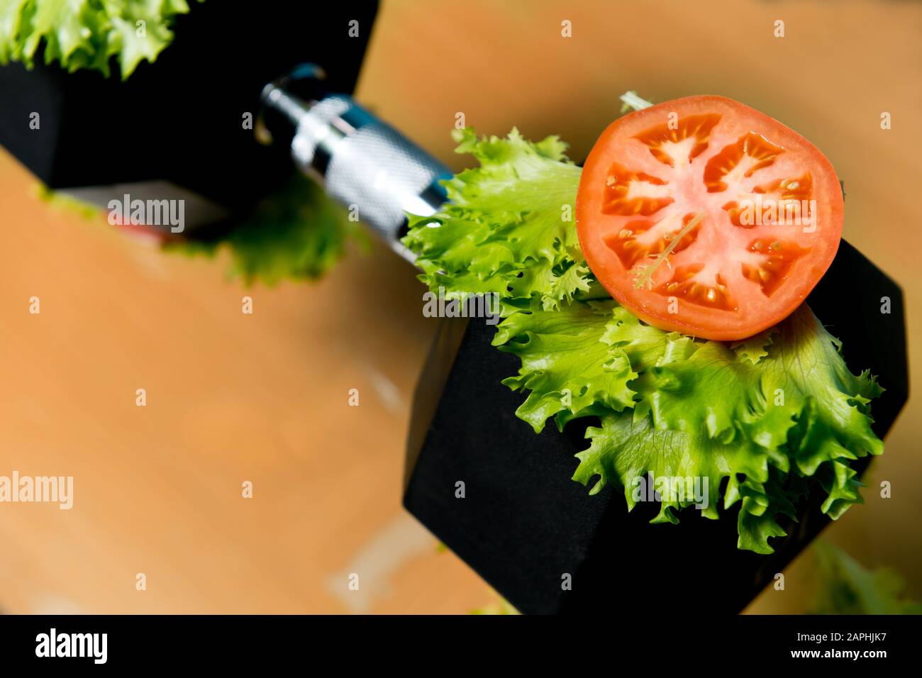 Eine Scheibe frische Tomate und Salat liegen auf einer großen Hanteln. Symbol für schwere Lebensmittel unter dem Deckmantel von frischem Gemüse. Schwere Lebensmittel Stockfoto