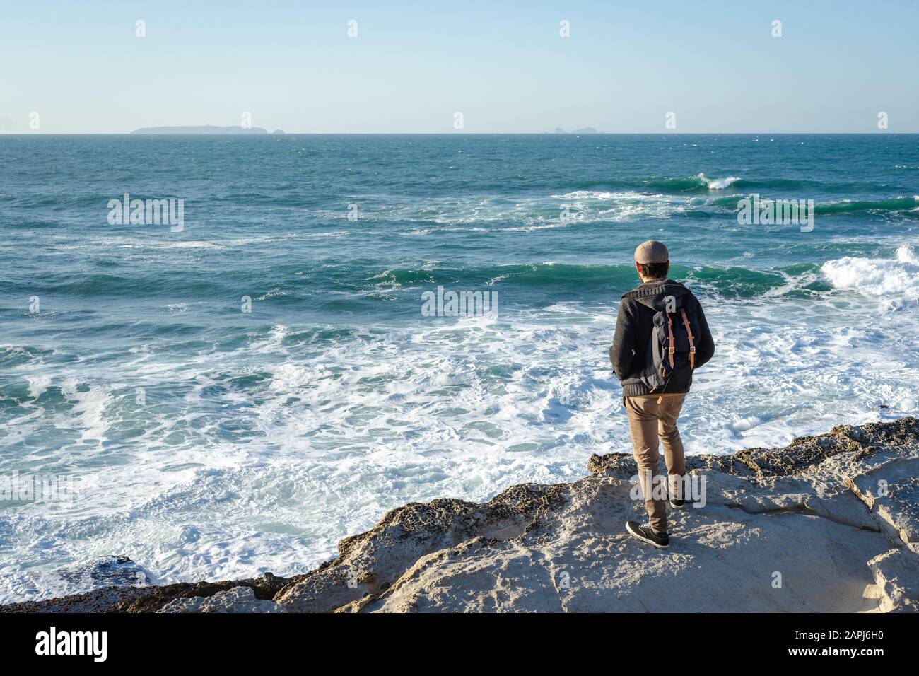 Person, die am Rand einer Felswand steht und die Wellen im Ozean betrachtet. Ansicht von hinten. Stockfoto