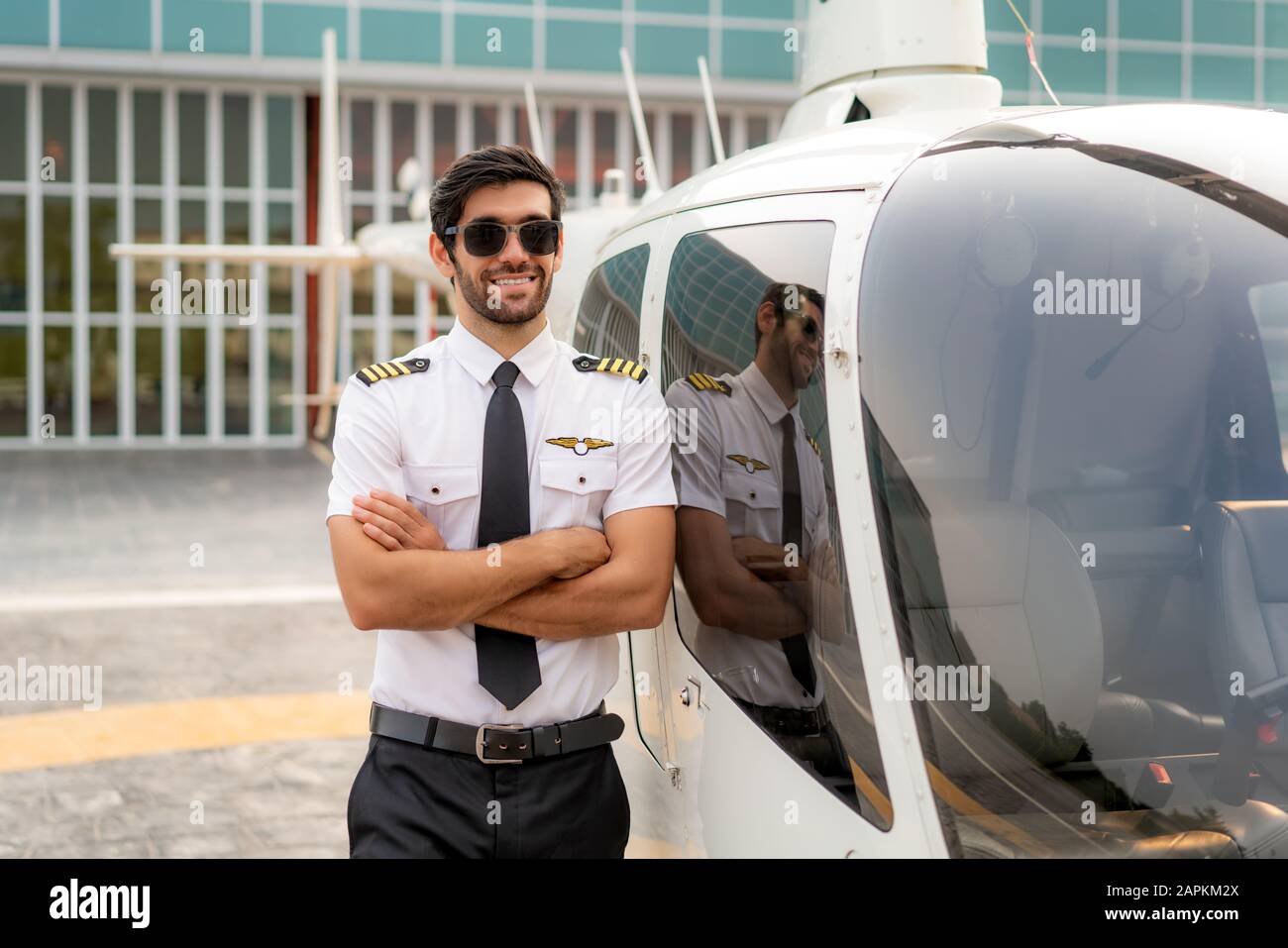 Portrait des gutaussehenden kommerziellen Piloten in Kapitänsuniform stehend und in der Nähe eines kleinen Privathubschraubers auf einem Landungspunkt mit Privatmann intelligent aussehend Stockfoto