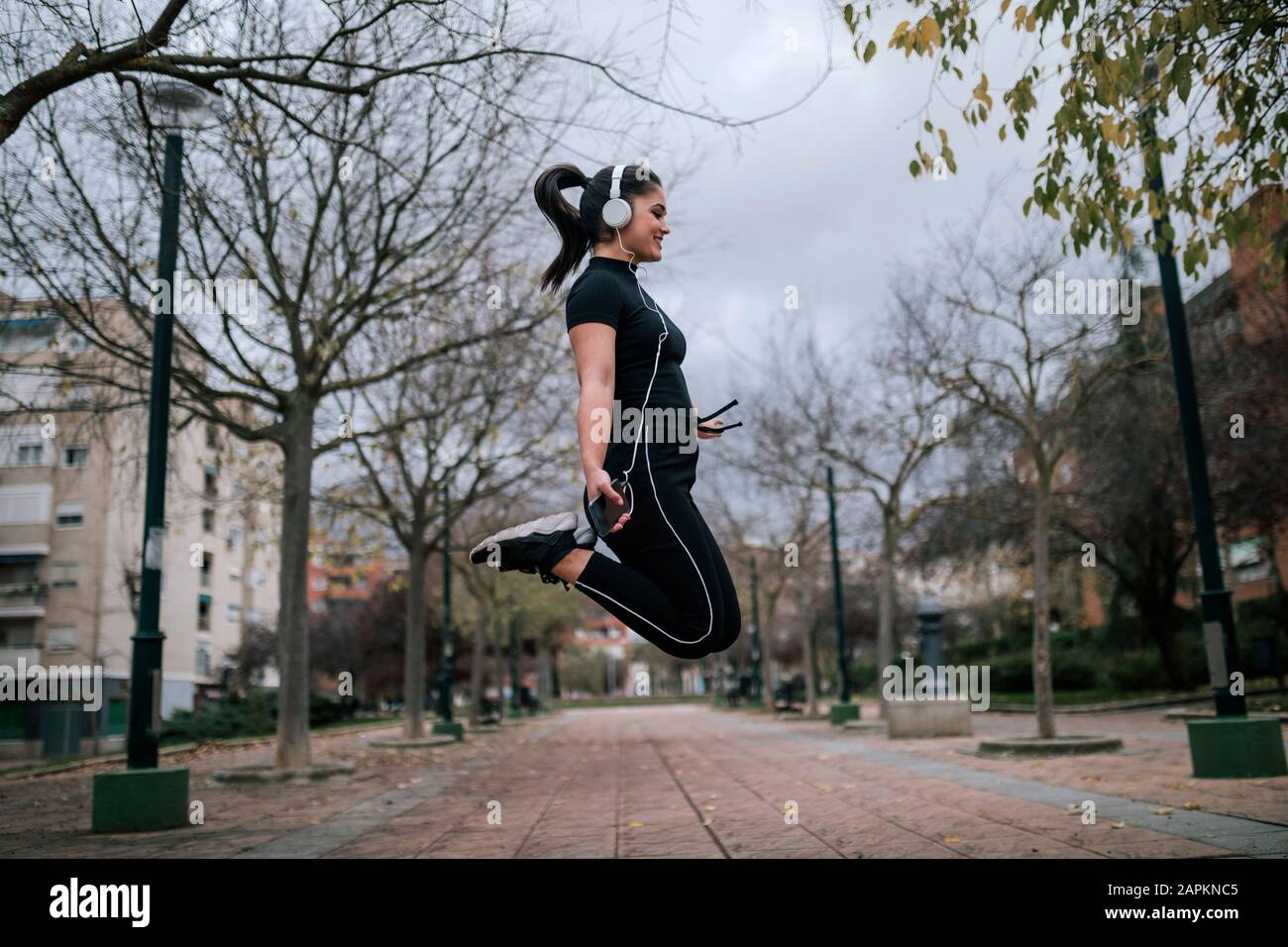 Junge Frau in schwarzer Sportswear springt in die Luft und hört Musik mit Kopfhörern Stockfoto
