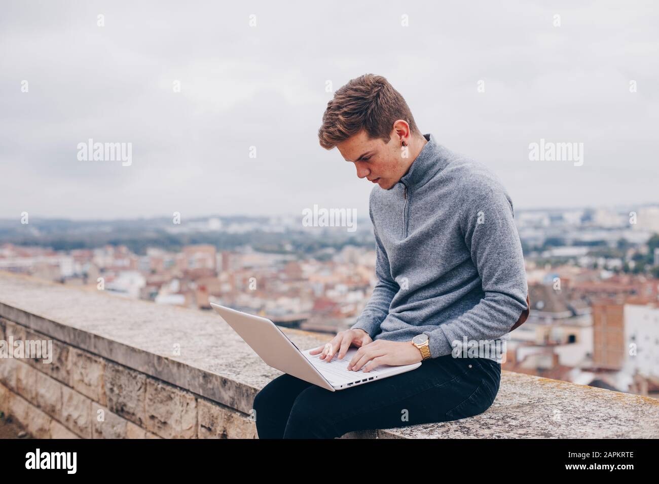 Blonder junger Mann sitzt an einer Wand und tippt auf seinem Laptop in Lleida Stockfoto