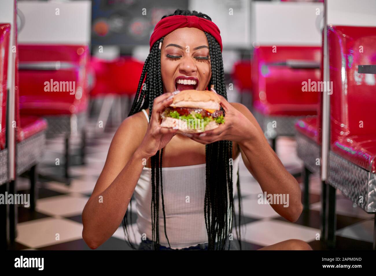 Junge Frau mit geflochtener Frisur auf dem Boden sitzend und einem großen Hamburger mit ihren beiden Händen zupft Stockfoto