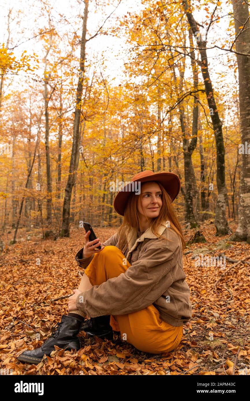 Porträt der modischen rotköpfigen jungen Frau mit Zellphoin, die im herbstlichen Wald herumsitzt Stockfoto