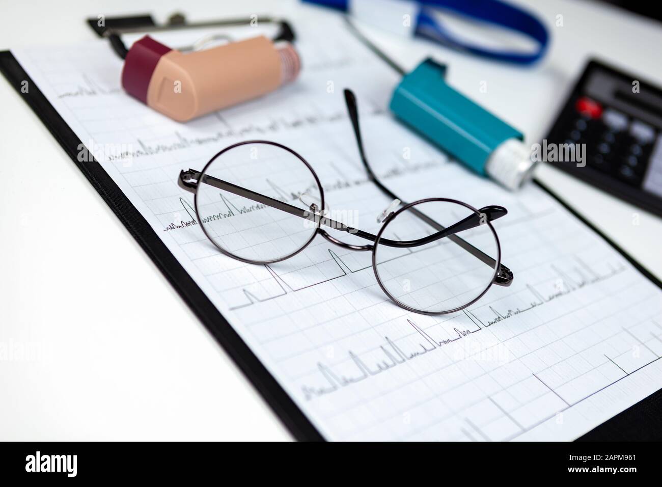 Ein Paar runde, randbeformte Brillen, die mit einigen Asthmainhalatoren und einer Wellenform auf einer Zwischenablage liegen Stockfoto
