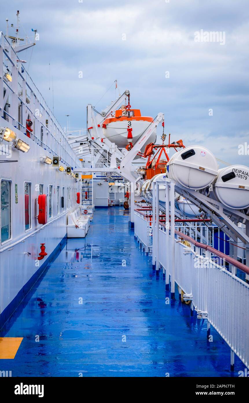 Offener Deckskorridor eines Fährschiffs mit Rettungsbooten und dem Hafen von Piräus im Hintergrund. Stockfoto