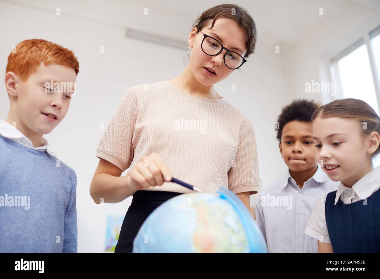 Junge Lehrerin in Brillen, die auf die Weltkugel zeigen und den Schulkindern Kontinente zeigen Stockfoto