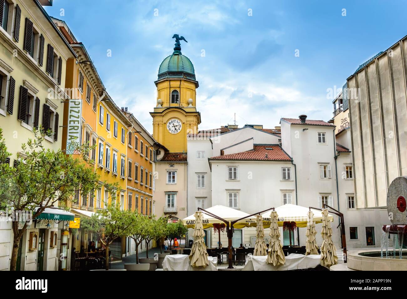 Rijeka, Kroatien - 19. Mai 2019: Platz hinter dem gelben City Clock Tower mit Café-Tischen, Sonnenschirmen und Olivenbäumen an einem sonnigen Tag. Gelbe Gebäude mit Witz Stockfoto