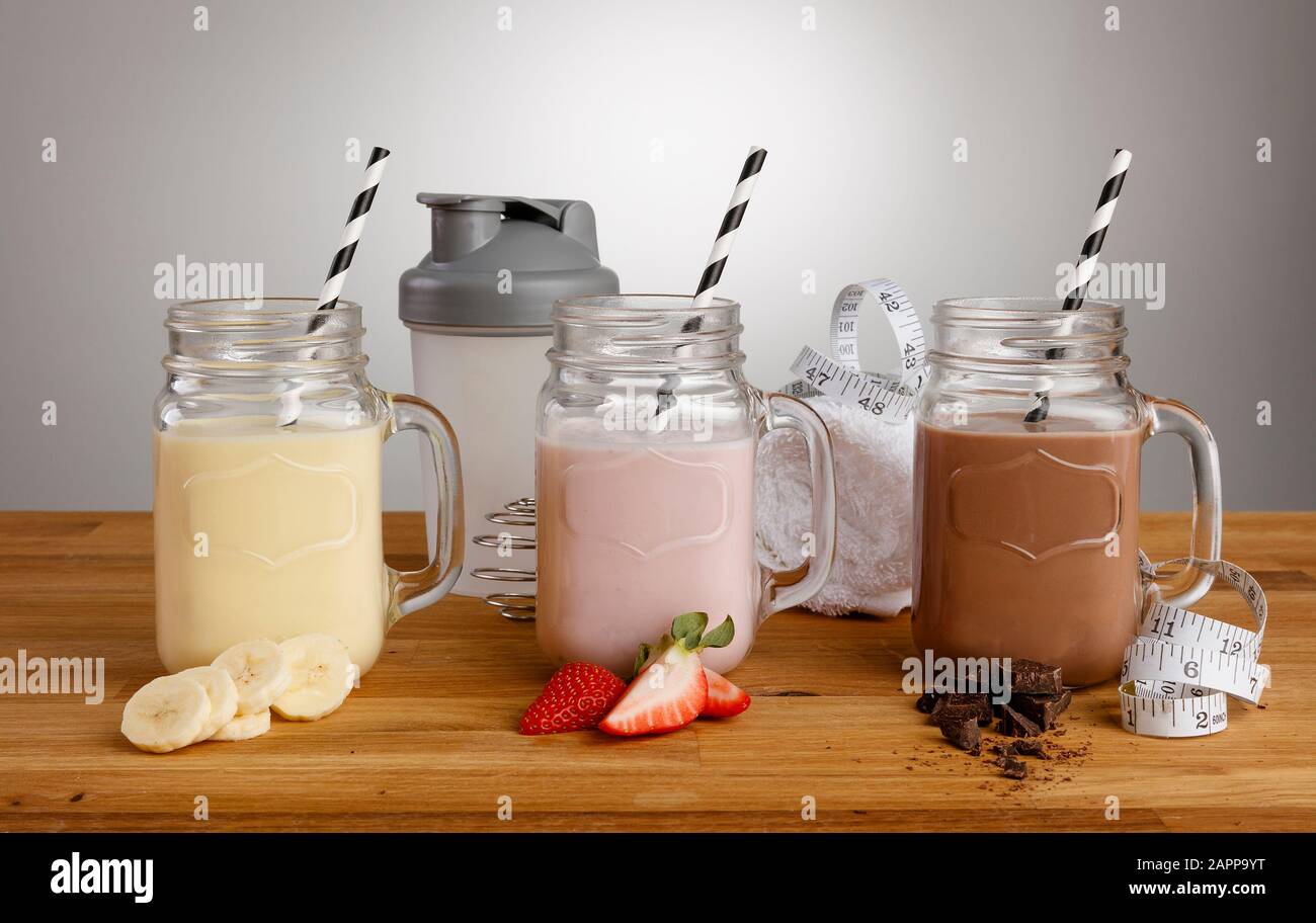 Banane, Erdbeere und Schokolade Milchshakes in Mason Glas Brille, mit Papierstraten, Zutaten und Shaker auf einem Holzarbeitspapier Stockfoto