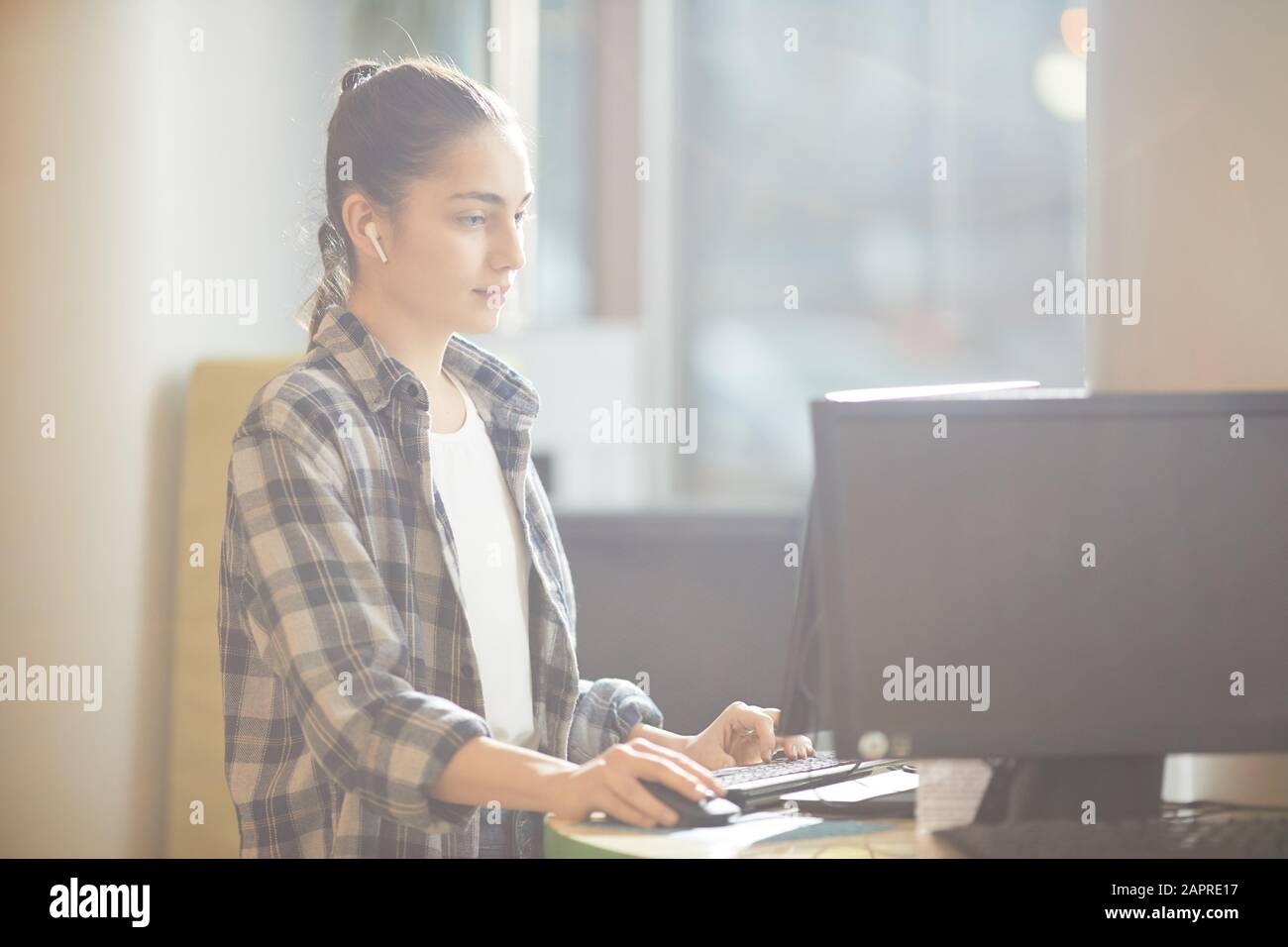 Junge Frau in legerer Kleidung sitzt am Tisch vor dem Computerbildschirm und tippt im Büro auf der Computertastatur Stockfoto