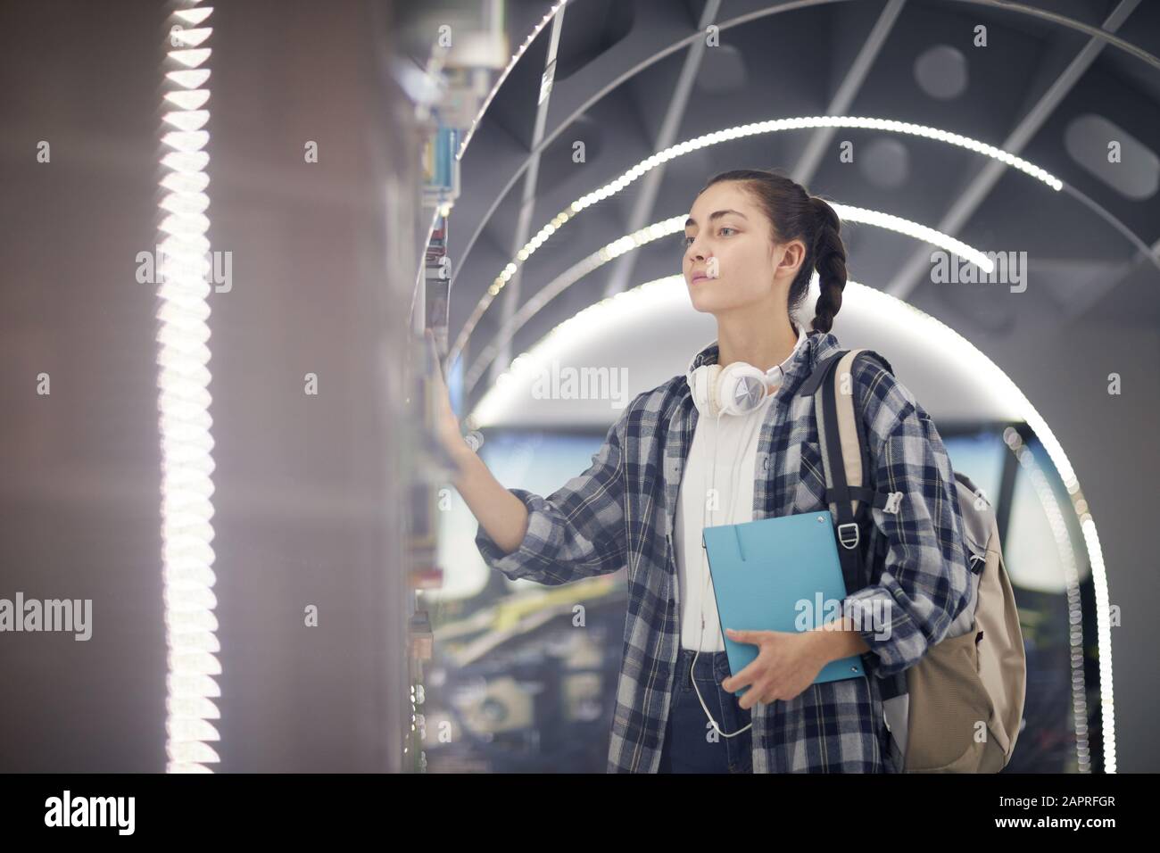 Junge Frau in legerer Kleidung mit Rucksack, die Buch aus dem Regal in der modernen Bibliothek mitnimmt Stockfoto