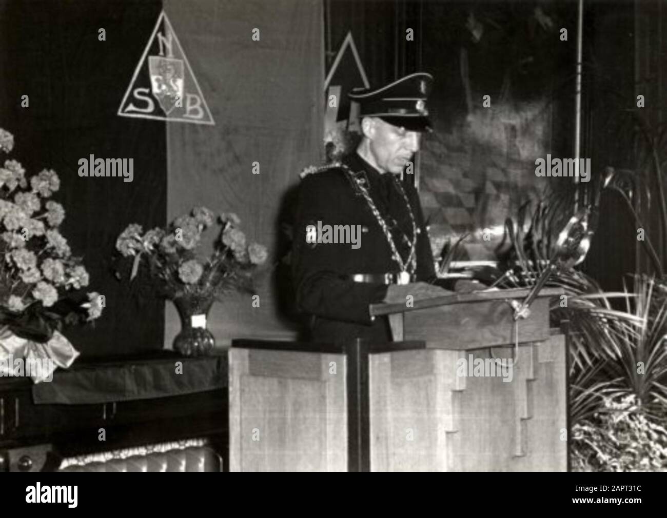 Der NSber Bürgermeister Jonkheer E.F. Sandberg steht hinter einer Katheder in SS-Uniform und liest seine Rede während seiner Installation als Bürgermeister von Kampen. Auf der Hülse das SS-Schild. Er trägt auch seine Amtskette. Hinter ihm steht das Emblem des NSB. Niederlande, 30. Mai 1941. Stockfoto