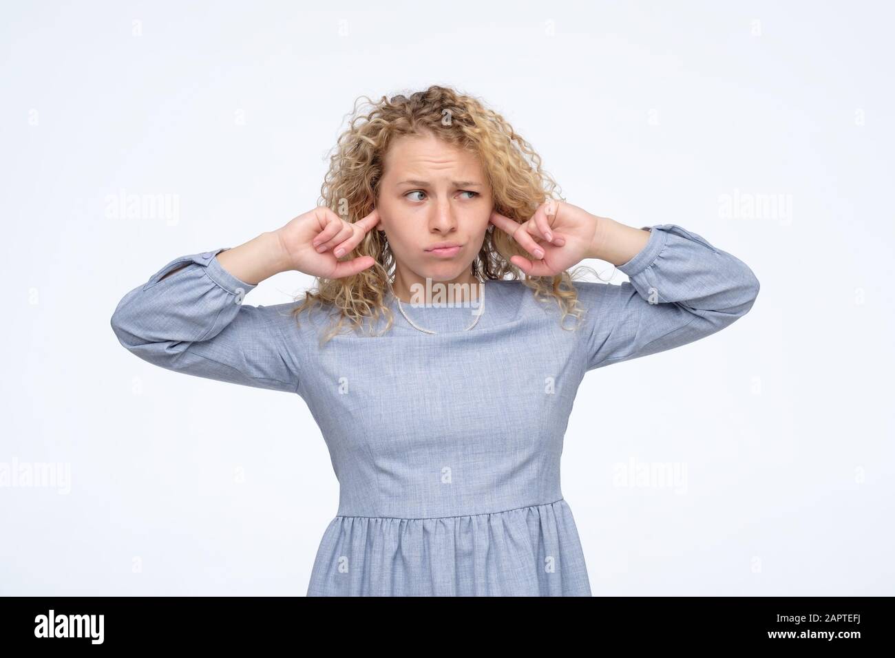 Junge blonde lockige Frau in blauem Kleid, die ihre Ohren bedeckt. Studio gedreht. Stockfoto