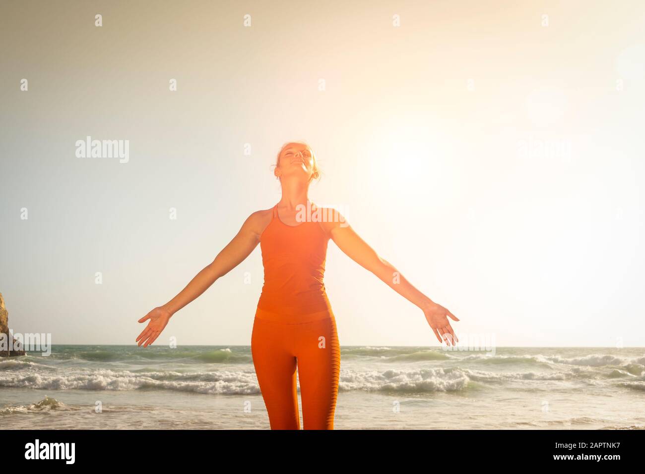 Fit Frau mit orangefarbener Sportbekleidung, die am Meer steht, mit den Armen und dem Gesicht zur Sonne Stockfoto