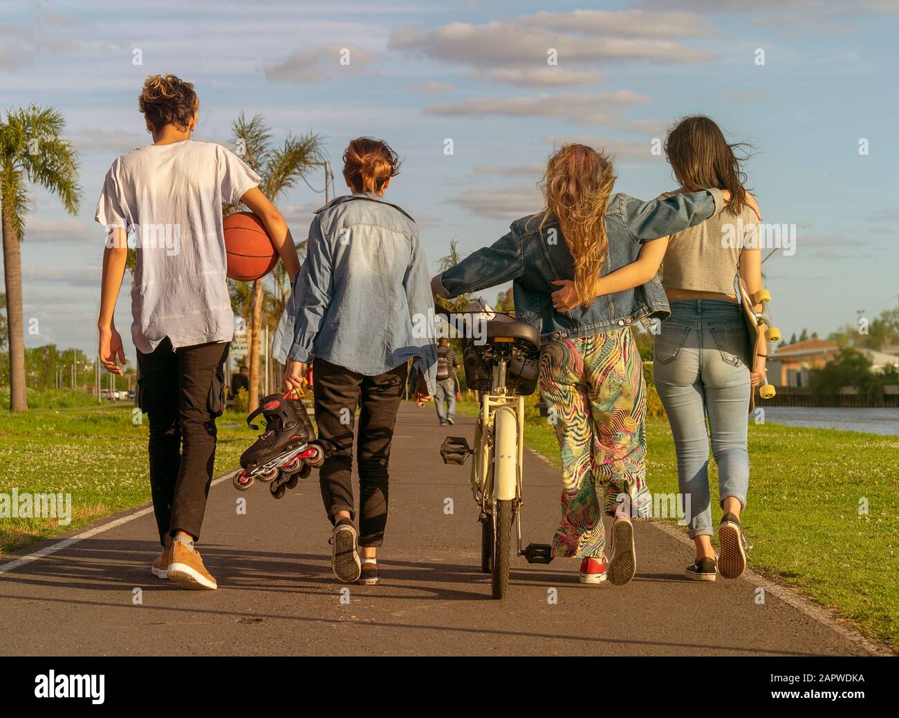 Porträt von hinter vier Jugendlichen, die auf einem Weg in einem Park mit einem Fahrrad, einem Skateboard, Rollskates und einem Basketball spazieren gehen Stockfoto