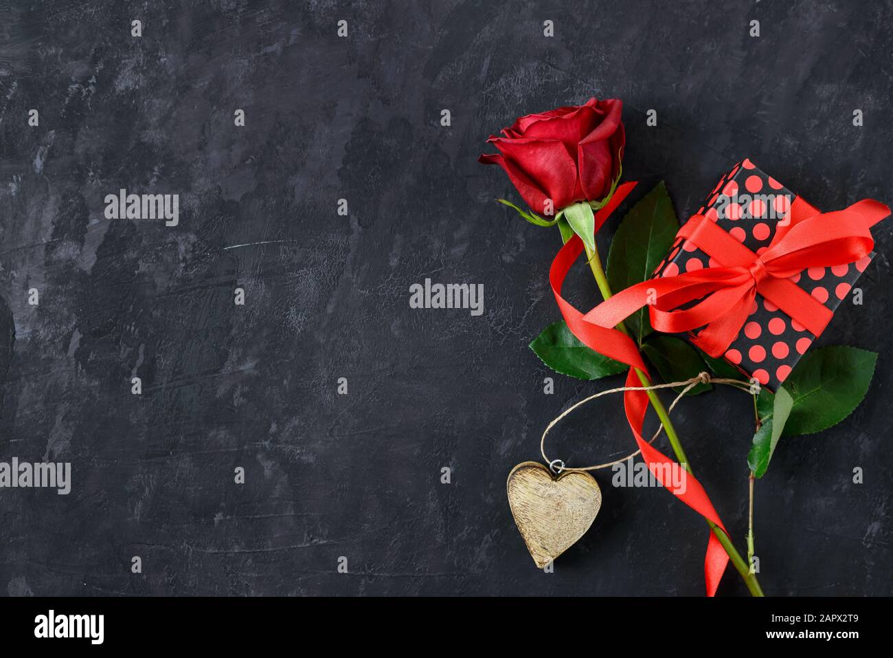 Grußkarte für Valentinstag mit romantischer roter Rose, Holzherz und Geschenkbox auf dunklem Tafel mit Platz für Text. Stockfoto