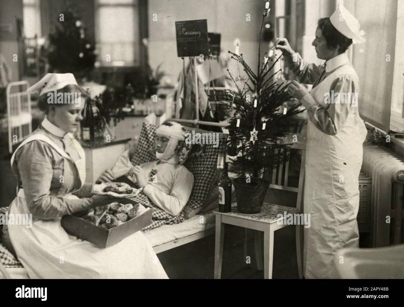 Ersten Weltkrieg Weihnachten in einem deutschen Krankenhaus für Kriegsverwundete. Ein verwundeter Soldat in einem Bett. Eine Krankenschwester schmückt einen kleinen Kirschbaum, eine andere sitzt am Rand des Bettes und bietet ihm etwas leckeres. Berlin, 1914. Stockfoto Ersten Weltkrieg Weihnachten in einem deutschen Krankenhaus für Kriegsverwundete. Ein verwundeter Soldat in einem Bett. Eine Krankenschwester schmückt einen kleinen Kirschbaum, eine andere sitzt am Rand des Bettes und bietet ihm etwas leckeres. Berlin, 1914. Stockfoto
