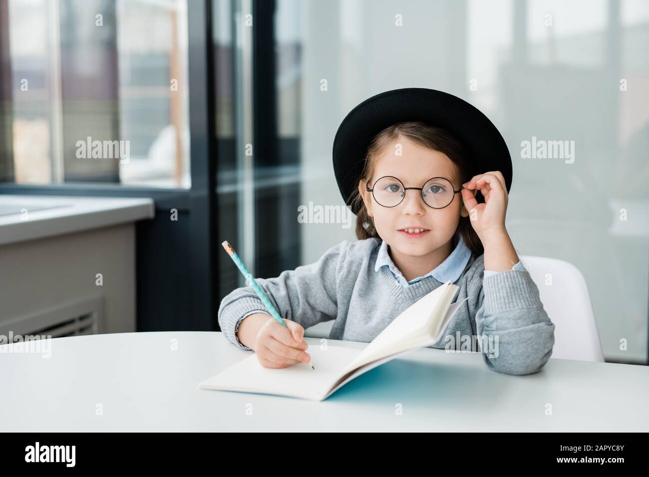 Liebenswürdige, clevere Schulmädchen im Hut, die Sie beim Erstellen von Notizen in einem Werbebuch betrachtet Stockfoto