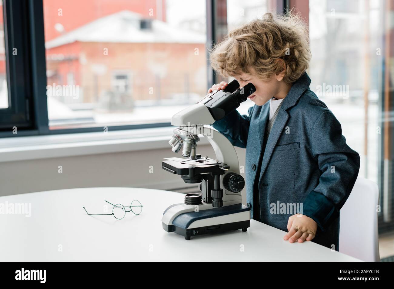 Cleverer Schuljunge mit geschweiften Haaren, die sich über den Schreibtisch beugen, während sie im Mikroskop schauen Stockfoto