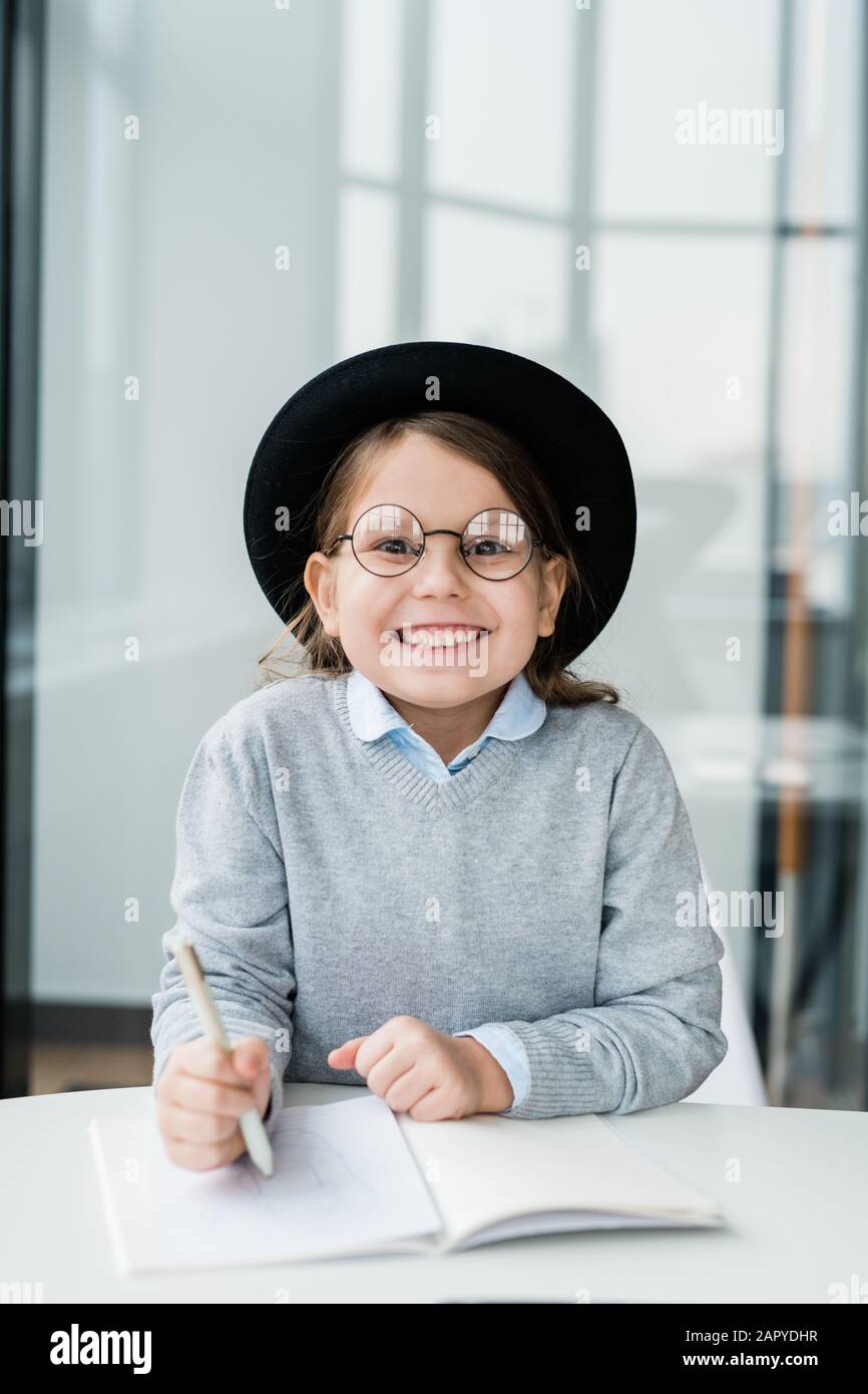 Fröhliches kleines Mädchen in Hut und Brille, das Notizen in einem Werbebuch macht Stockfoto