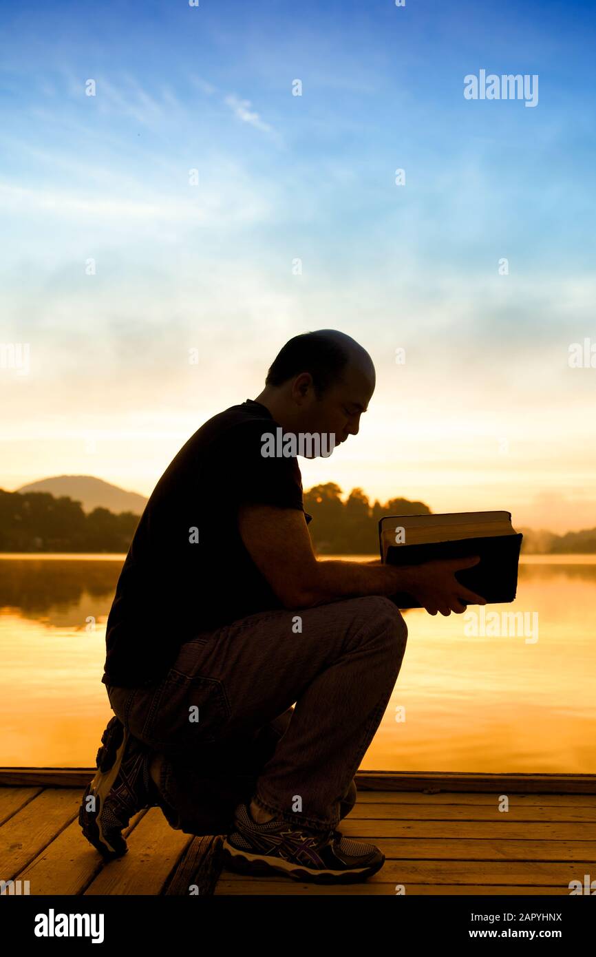 Mann liest ein Buch auf einer Holzbrücke umgeben von Der See und die Hügel während des Sonnenuntergangs Stockfoto