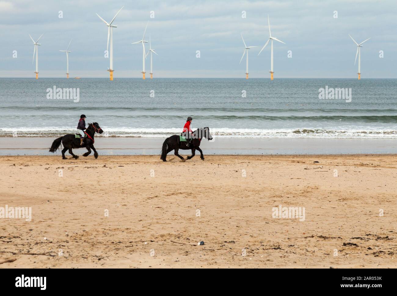 Zwei Frauen, die Pferde am Redcar Strand mit den Windenergieanlagen im Hintergrund im Nordosten Englands, Großbritannien, reiten Stockfoto