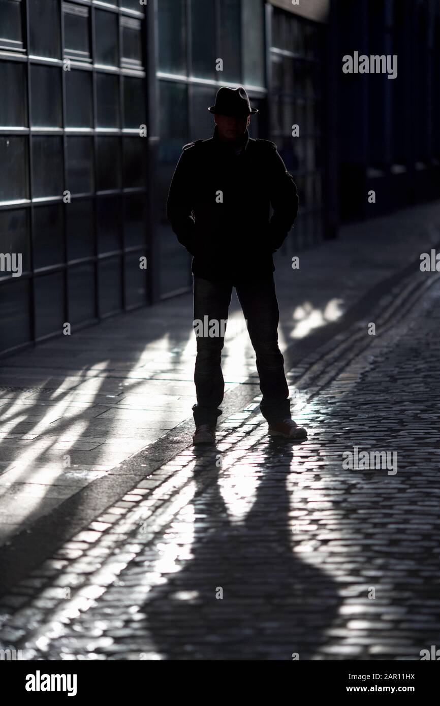 Geheimnisvoller Mann mit Hut und Mantel, der im Sonnenlicht auf einer alten gepflasterten Straße silhouettiert steht Stockfoto