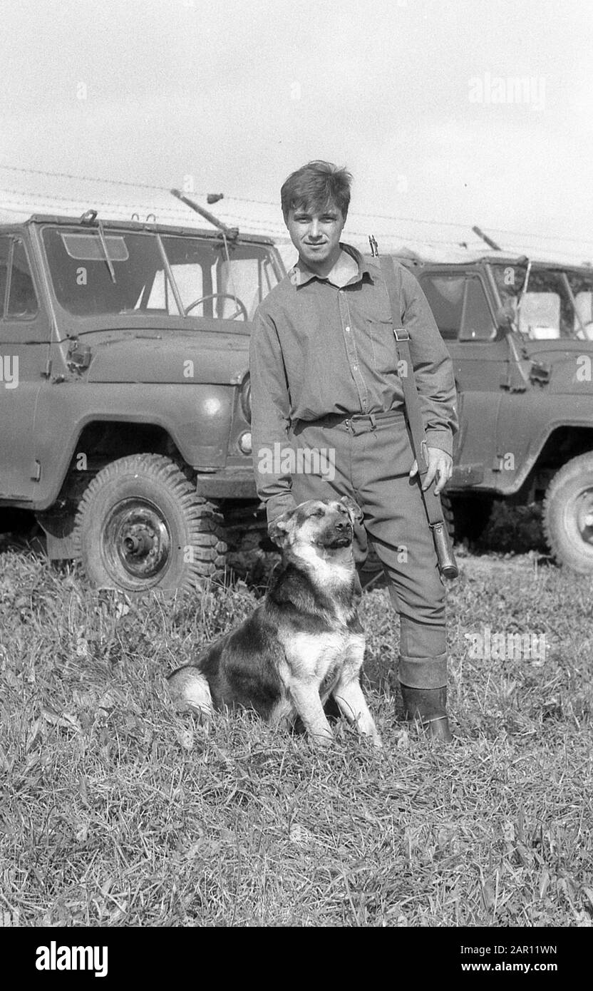 STUPINO, REGION MOSKAU, RUSSLAND - CIRCA 1993: Porträt des Soldaten der russischen Armee mit einem Hund (Hirte) im Hintergrund-SUV UAZ. Schwarz und Weiß. Filmscan. Große Körnung. Stockfoto