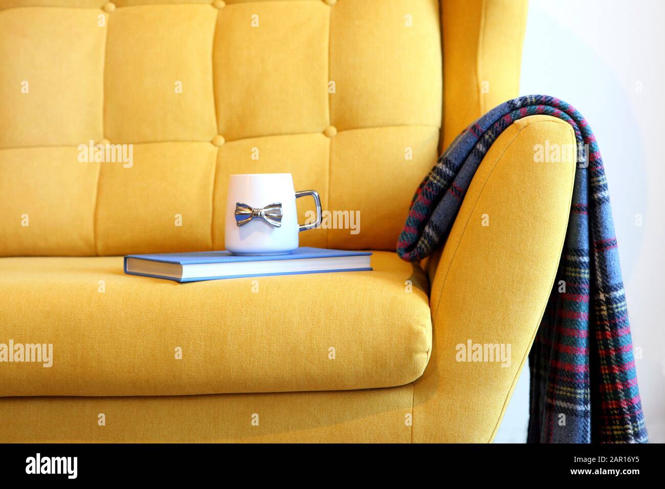 Tasse Tee und blaues Buch auf gelbem Trainer mit Decke. Details zum Stillleben im Inneren des Wohnzimmers. Gemütliches Innenleben, Hauskomfort Stockfoto