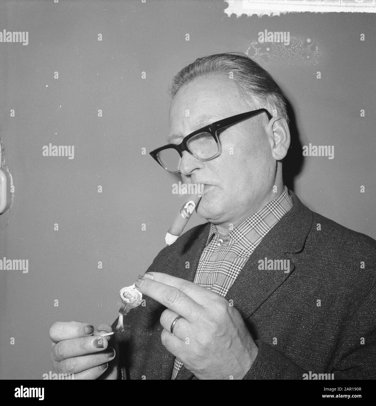 Große Männer und Tyrannen, Zigarrenriemen, der Mann legt ein Band mit dem Porträt von Hitler in Brand Anmerkung: Um die Zigarre ist ein Zigarrenriemen mit einem Bild von Präsident Kennedy Datum: 2. Dezember 1964 Schlüsselwörter: Rauchen, Zigarren, Zigarrenbänder Stockfoto