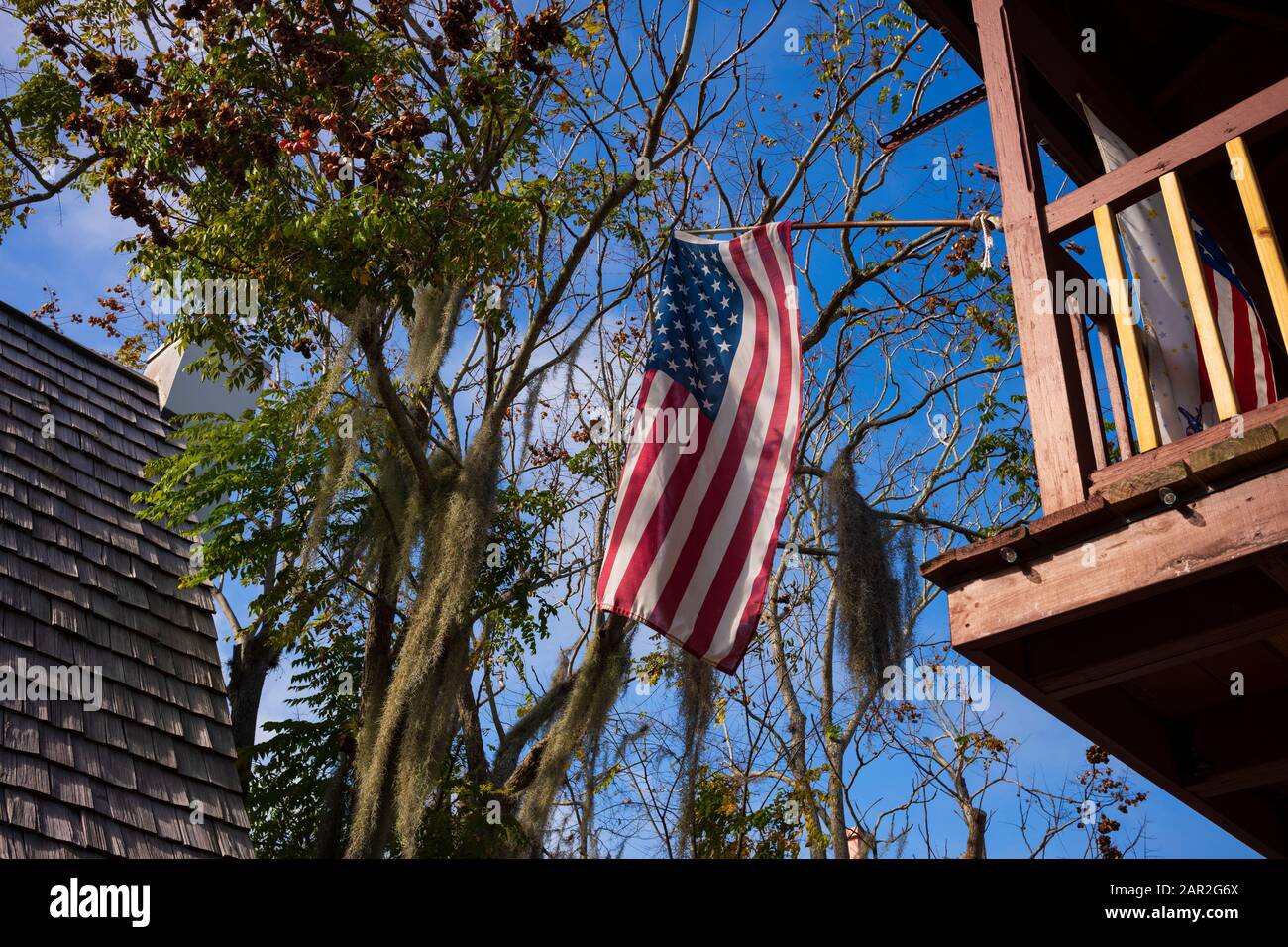 Die amerikanische Flagge fliegt hoch von einem Balkon mit einem Baum mit hängendem spanischem Moos, blauem Himmel und einem Haus mit Zedernschindeln im Hintergrund. Stockfoto